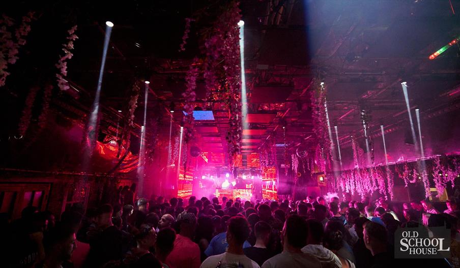 A crowded nightclub filled with people facing a brightly lit stage, with beams of white and colored lights shining down and floral decorations hanging
