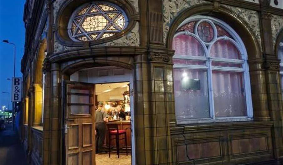A nighttime street view of the ornate exterior of Hotel Alexandra, with a warmly lit entrance showing people inside at the bar. The building features