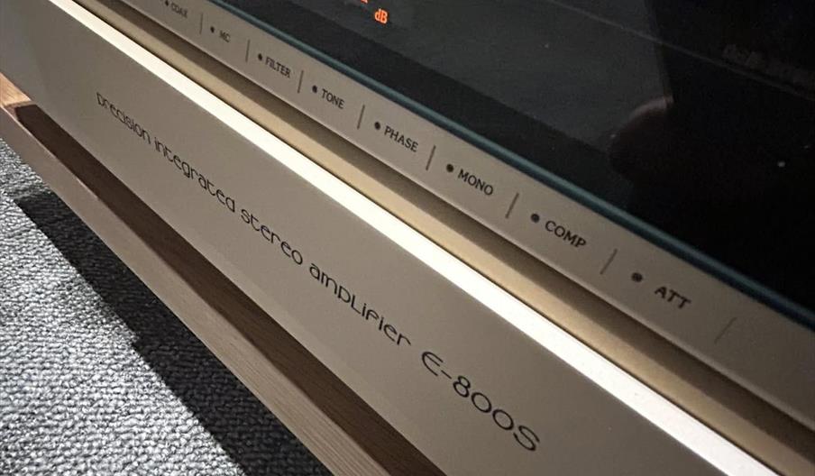 Close-up of a stereo amplifier display showing analog dials with "dB Peak Power Level" and an illuminated digital readout of "-42.5 dB"; the amplifier