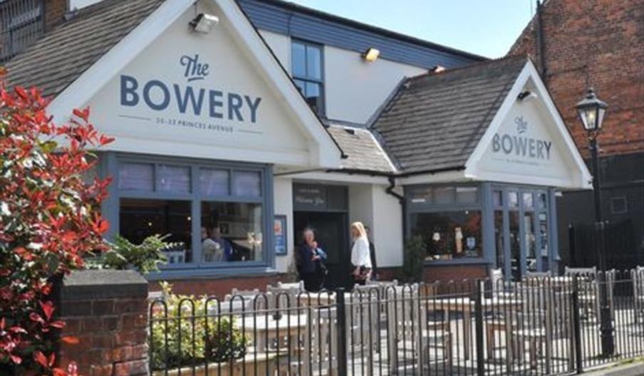 A street view of "The Bowery," a light-colored building with large front windows and two peaked roof sections displaying its name. There are outdoor t
