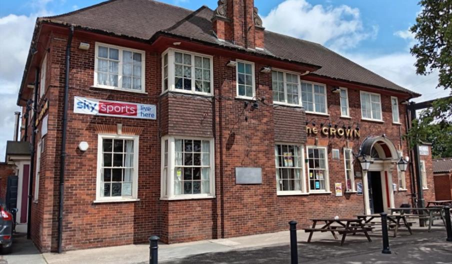A two-story red brick pub called "The Crown" with large white-framed windows and several signs, including one with a stylized crown logo above the ent