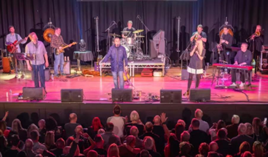 A band performs on a well-lit stage with multiple musicians playing instruments including guitar, drums, keyboard, and brass, while three vocalists st