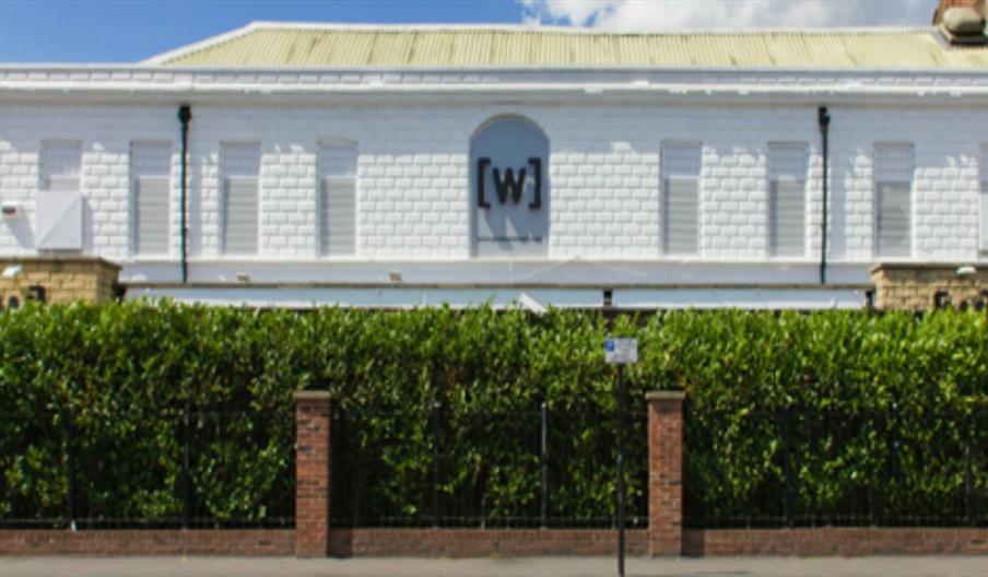 A white brick building, the Welly Club with a sign featuring a bold black “[w]” on the front, partially obscured by a tall green hedge and a brick-and