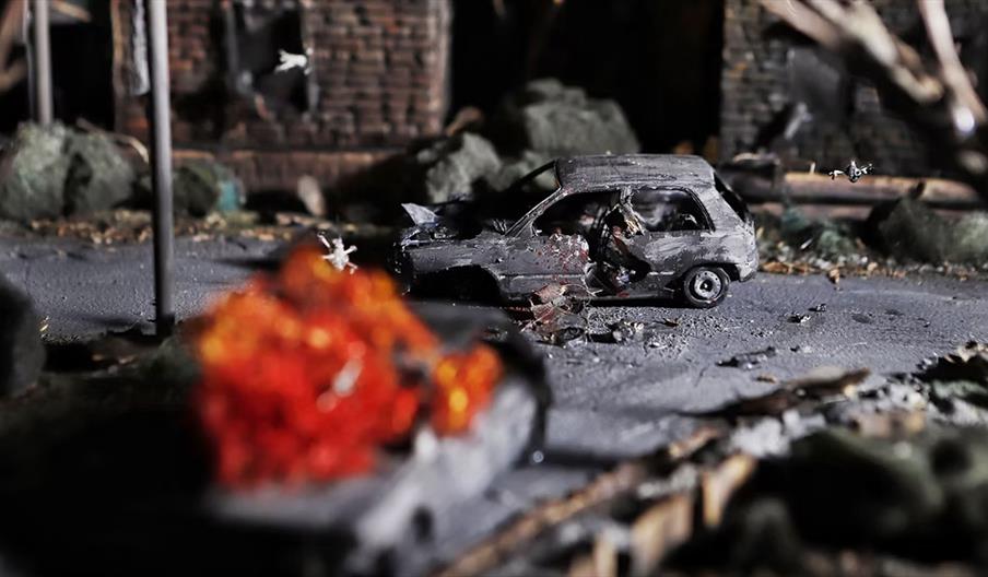 A damaged grey car sits on a debris-strewn road with a large explosion and flames in the foreground, suggesting a scene of destruction in an urban are