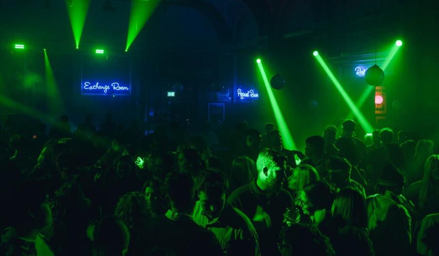 A crowded nightclub filled with people dancing under green stage lights, with neon signs reading "Exchange Room" and "Royal Room" visible on the back