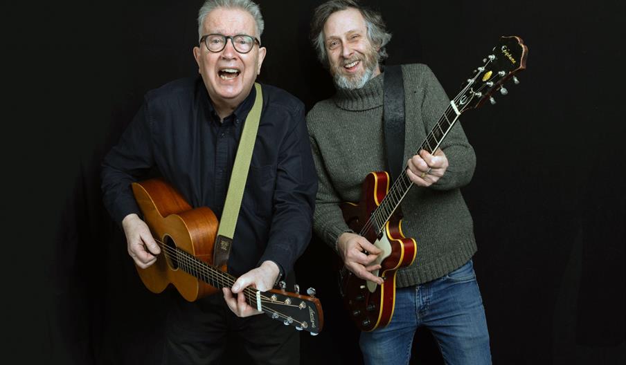 Two older men stand side by side against a black background, smiling and playing guitars; the man on the left holds an acoustic guitar and wears glass