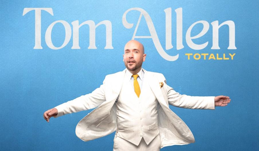 A person in a white suit and yellow tie stands with arms outstretched against a blue background with large text that reads "Tom Allen" and smaller yel