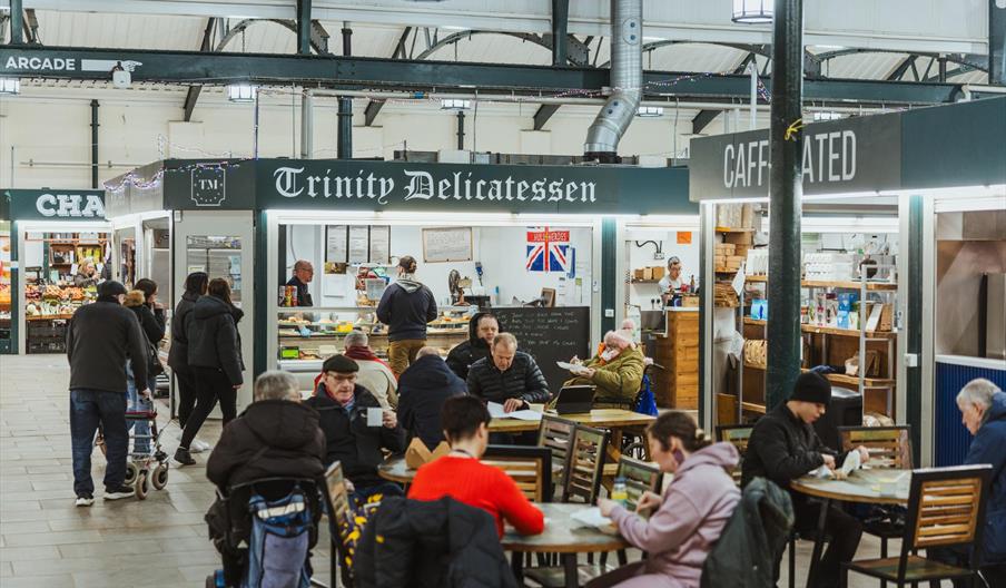 Indoor shot of Trinity market. Several table are scattered throughout the space with people sat on chairs around them. To the side sits the service ha