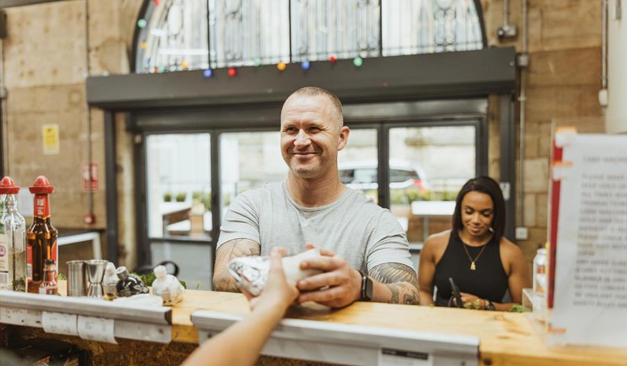A smiling man is served food over the counter in Trinity Market. Behind him stands a woman waiting to be served.