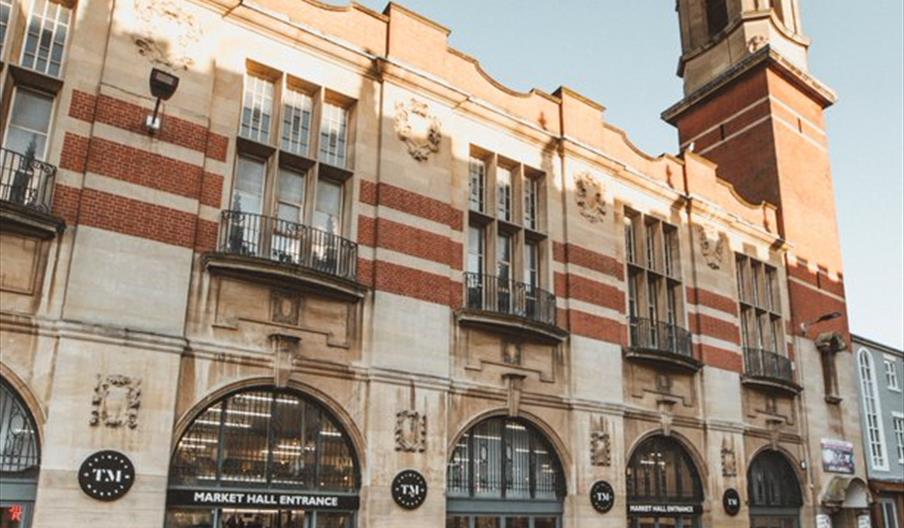 A historic Trinity Market building with arched windows and red-and-tan brickwork stands along a cobblestone street; signs above the main entrances rea