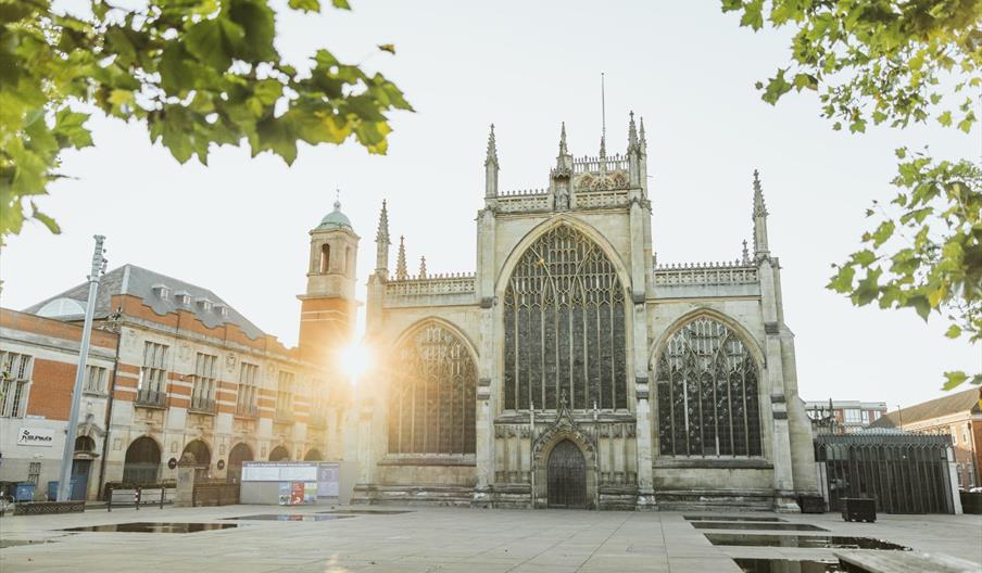 Hull Minster, a large Gothic-style church with tall arched windows and spires stands in an open square, with the sun shining through nearby buildings