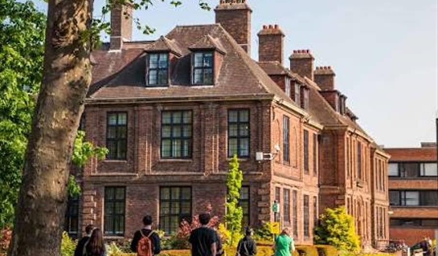 A group of students walks on a pathway toward a historic brick building on the University of Hull campus, with a sign in the foreground displaying the