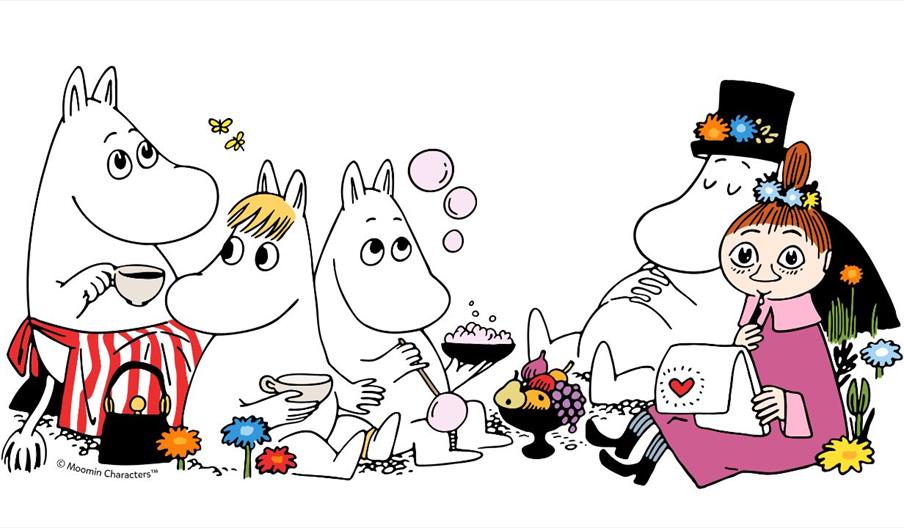 Five cartoon Moomin characters sit together having a picnic, surrounded by flowers, with tea, sweets, and a basket of fruit. One character wears a str
