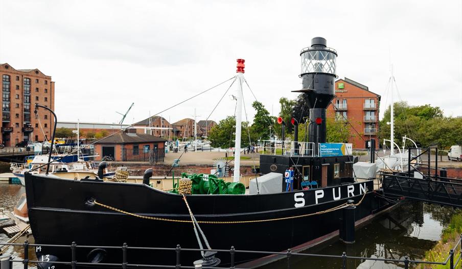 A large historic black ship, "SPURN" is docked in a harbor, featuring a prominent lighthouse-style tower on its deck. Surrounding the ship are other b