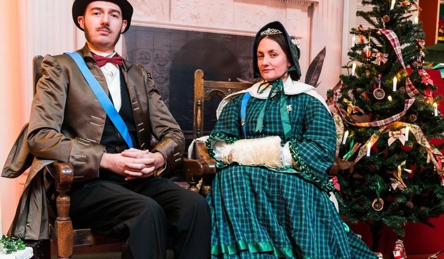 A man and a woman dressed in elaborate Victorian-era costumes sit side by side on wooden chairs. They are indoors next to a decorated Christmas tree,