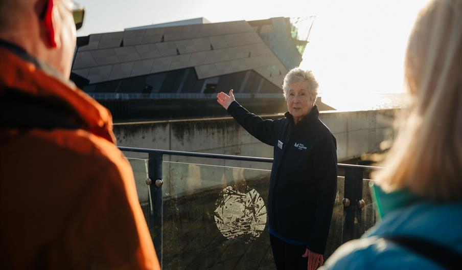 An older woman in a navy jacket gestures toward a modern, angular building while standing outside on a sunny day, with two people listening in the for