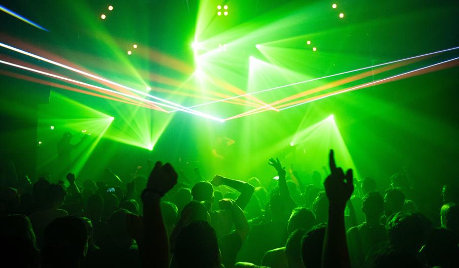 A crowd of people in the Waterfront Nightclub with raised hands, illuminated by vibrant green laser lights and beams cutting through the smoky, dark a