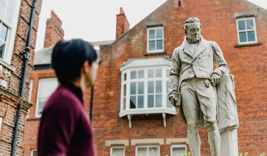 A person in a burgundy jacket stands in the foreground looking at a statue of William Wilberforce, which depicts him in formal 19th-century attire hol