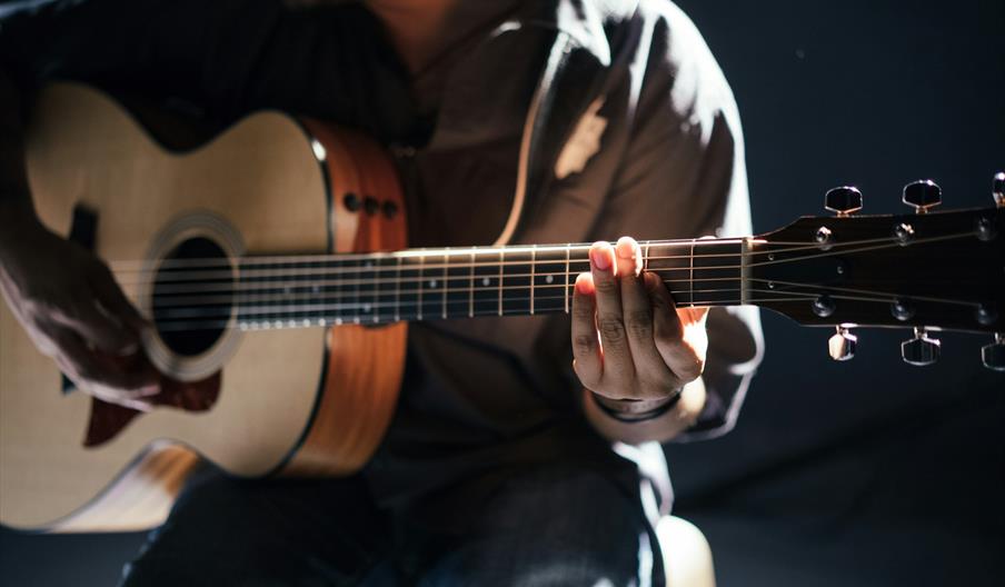 A person plays an acoustic guitar, with focus on their hands strumming and pressing the strings on the fretboard under dramatic lighting.