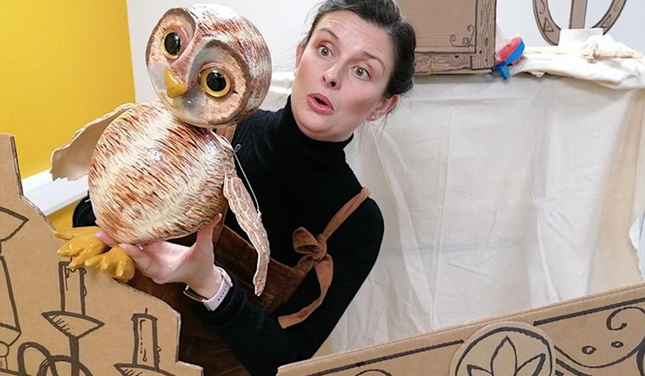 A woman wearing a black turtleneck and brown overalls holds a large owl puppet, looking surprised or animated. She stands amid cardboard props and set
