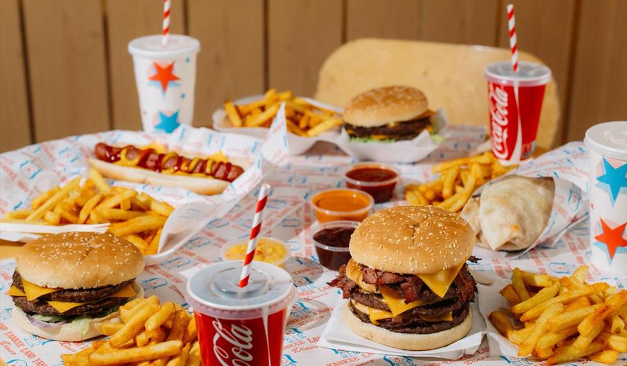 A table filled with classic fast food items including burgers, fries, a hot dog with ketchup and mustard, a wrapped burrito, several dipping sauces, a
