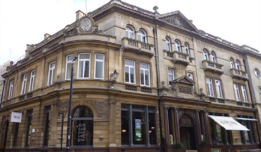 Street view of a Yates bar and restaurant with decorative exterior, large front windows, outdoor tables and chairs, and white awnings displaying the n