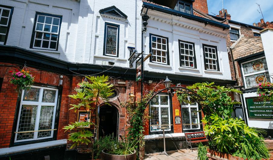 Exterior of Ye Olde White Hart, a traditional English pub with white and red brick walls, large multi-pane windows, and hanging flower baskets. The en