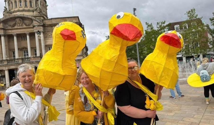 Three smiling women stand outdoors in a plaza, each holding a large, yellow duck-shaped puppet or lantern. They are surrounded by people and historic
