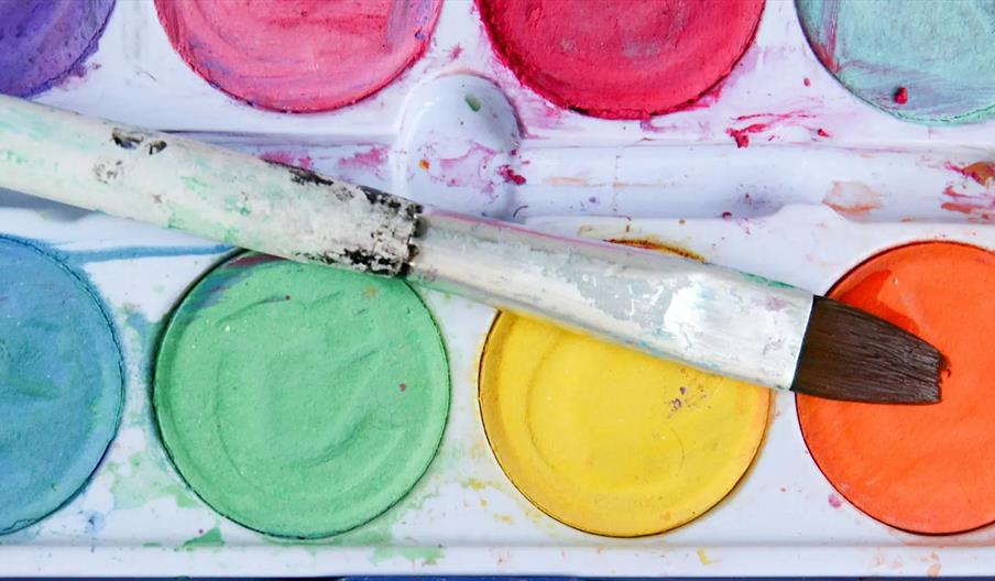 A close-up of a watercolor paint palette with various colors, including blue, green, yellow, and orange, with a paintbrush resting diagonally across t