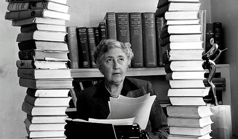 Agatha Christie, an older woman sits at a desk with a typewriter, holding papers, surrounded by two towering stacks of books on either side. Bookshelv