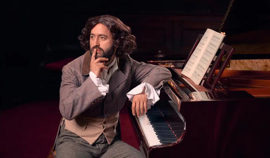 A man in historical clothing with long, wavy hair sits at a piano, resting one hand on the keys and the other on his chin in a thoughtful pose; sheet