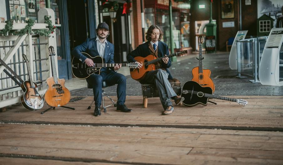 Two men in suits sit on a wooden platform, each playing an acoustic guitar, with four additional guitars resting on stands or the ground around them i