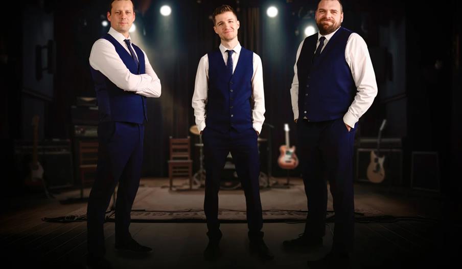 Three men dressed in matching navy blue vests and ties stand confidently on a stage, with musical instruments and stage lights in the background.