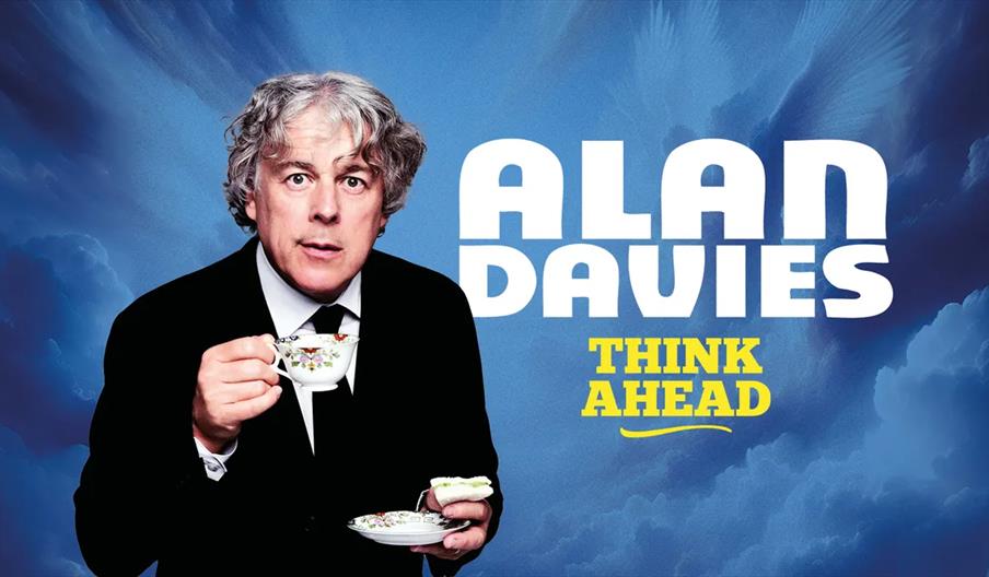 Alan Davies with curly gray hair, dressed in a black suit and tie, holds a teacup and saucer against a blue cloudy background. Large white and yellow