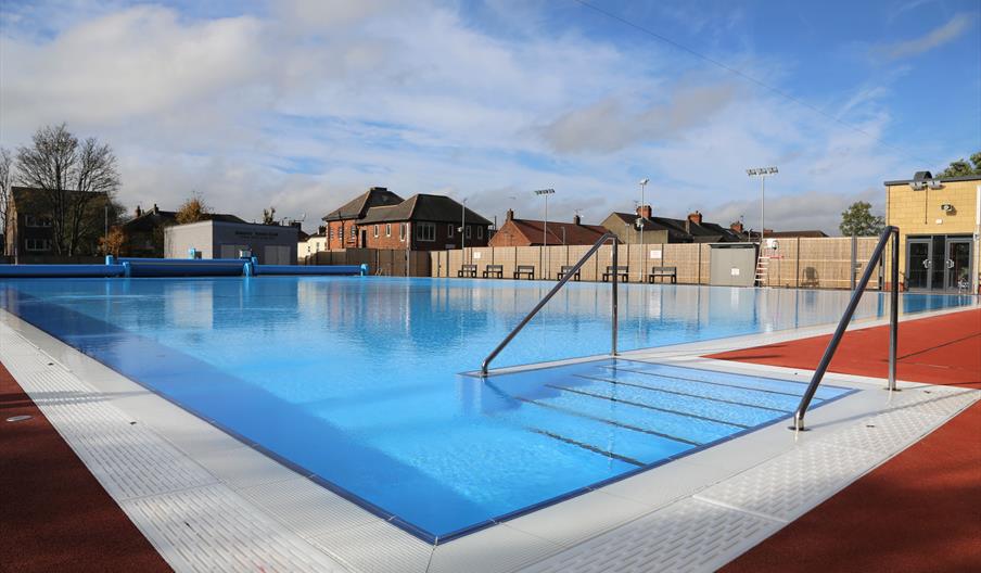 An outdoor swimming pool with clear blue water and metal handrails at the entrance, surrounded by red non-slip flooring and with buildings and trees v