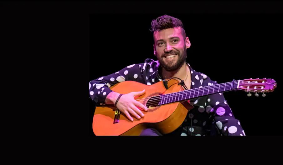 A smiling man with a beard, wearing a black shirt with large white polka dots, holds an acoustic guitar while seated against a black background.
