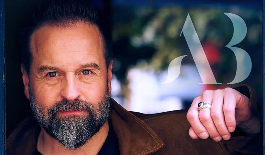 Alfie Boe stands with a neatly groomed beard and mustache, wearing a brown jacket, by a window with his hand resting on the frame; a stylized "AB" log