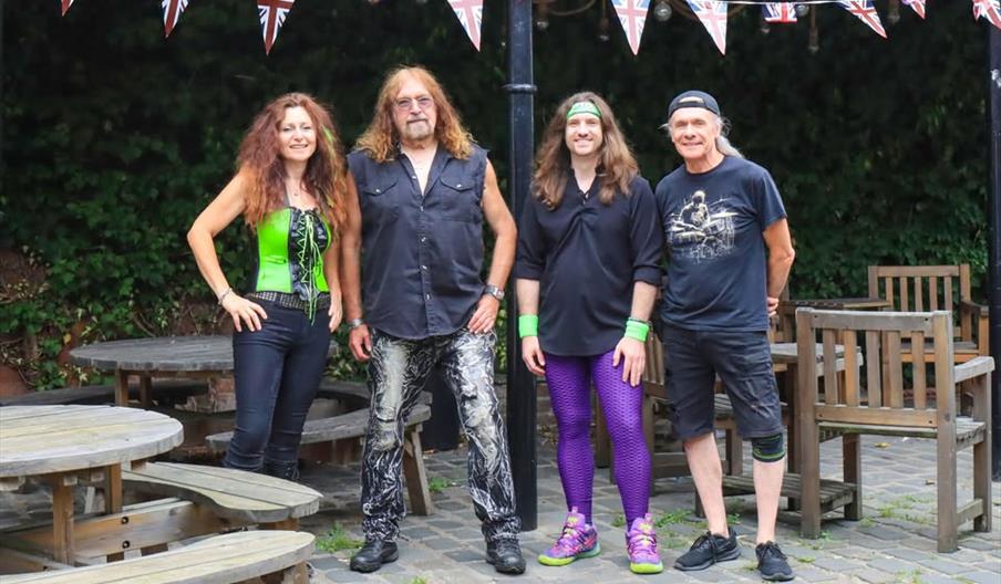 Four people stand outdoors in front of picnic tables and wooden chairs, with Union Jack bunting hanging above. The group, dressed in colorful and casu