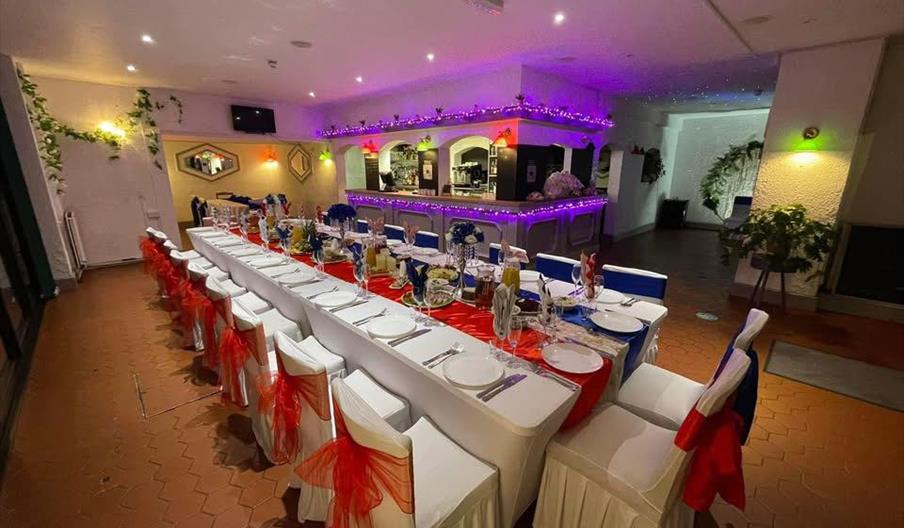A long decorated table, with colourful sashes on the back, with a bar behind decorated with purple fairylights.