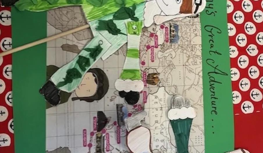 A handmade craft display shows a map with cartoon-style paper figures of explorers in green and white outfits. The background features green and red p