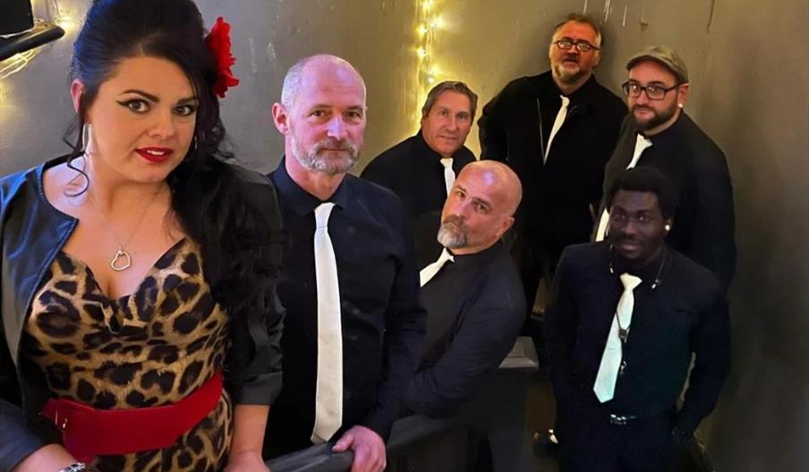 A group of seven adults, six men and one woman, stand on a staircase decorated with string lights. The woman in front wears a leopard print dress with