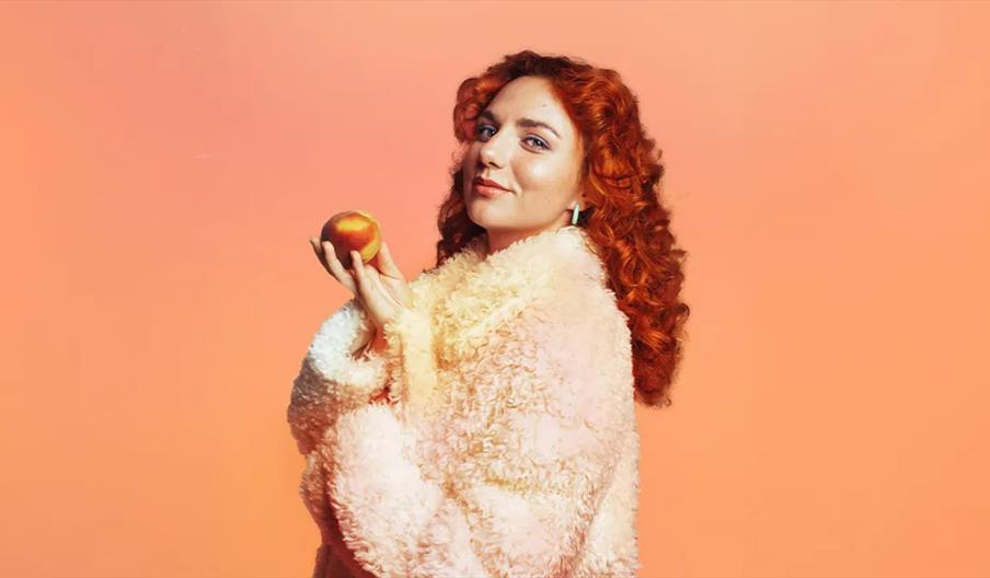 Ania Magliano with curly red hair wearing a fluffy, pale pink coat holds a peach in one hand, posing against a peach-colored background.