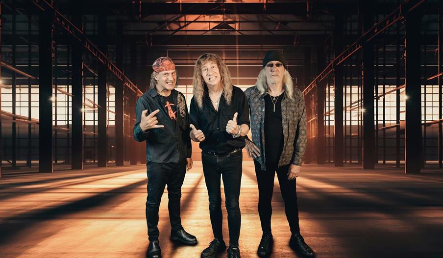 Three men with long hair, dressed in black clothing and standing confidently, pose inside an industrial warehouse with warm lighting and metal beams i