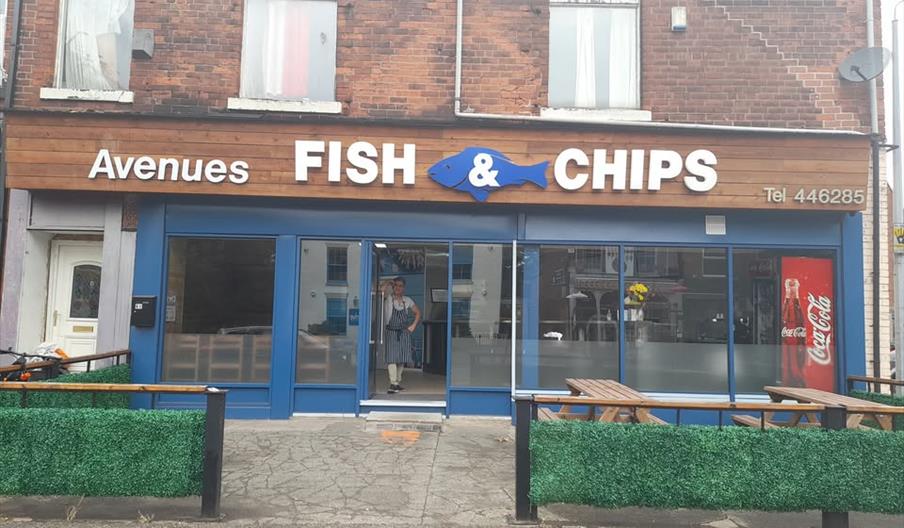 The front of Avenues Fish and Chips, picnic benches stand outside and barriers covered in fake greenery, whilst the owner casually stands in the doorw