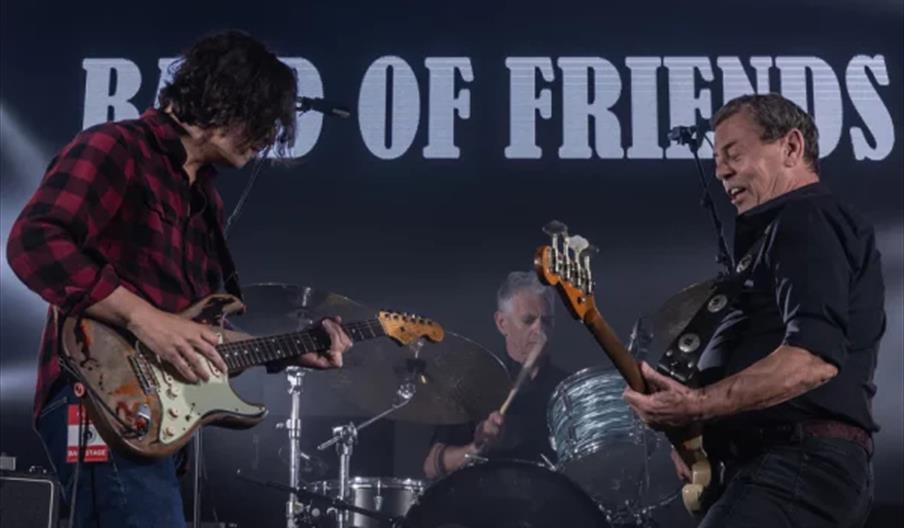 Three musicians perform energetically on stage, with two guitarists at the front and a drummer in the background. The words "BAND OF FRIENDS" are part