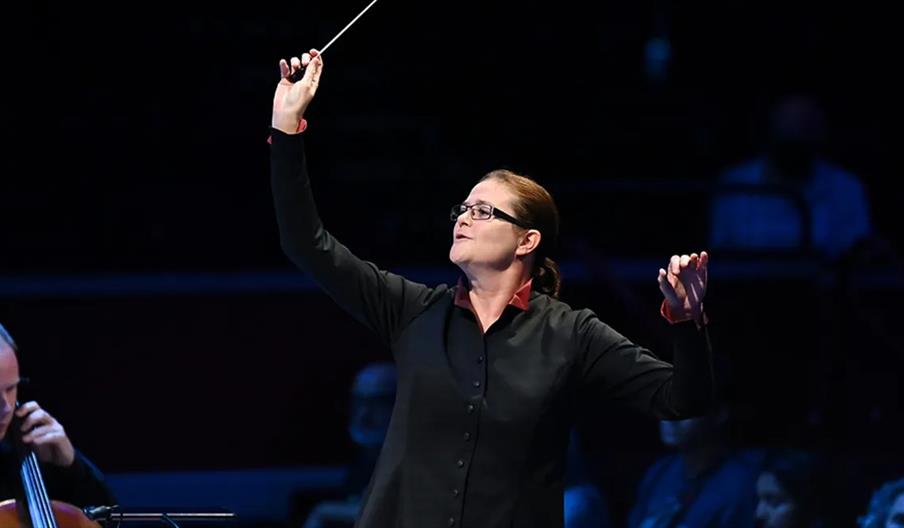 A conductor stands on stage, holding a baton in one hand and raising the other, leading an orchestra with focused expression while wearing glasses and
