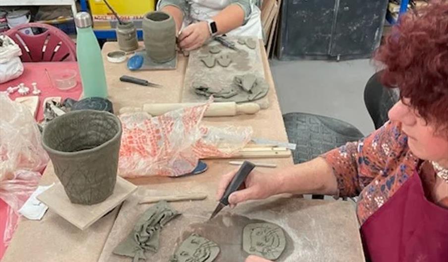 Two women sit at a table in a pottery studio, shaping and decorating pieces of clay. Various clay tools, unfinished pottery, and sculpted objects are