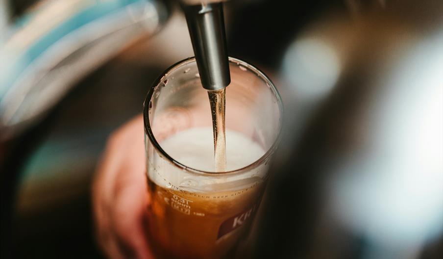 A close-up of a beer tap pouring beer into a clear glass, with foam forming at the top and a hand holding the glass.