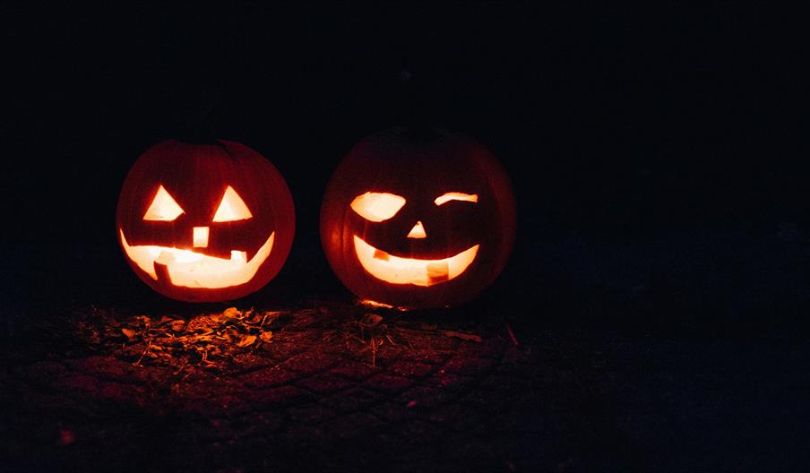 Two carved pumpkins with glowing faces sit side by side in the dark; the one on the left has a classic jagged smile and triangular eyes, while the one