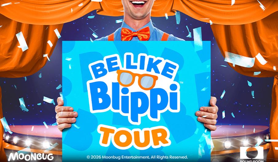 A person dressed in a blue shirt with an orange bow tie and suspenders holds a large sign that reads "Be Like Blippi Tour" with orange glasses in the